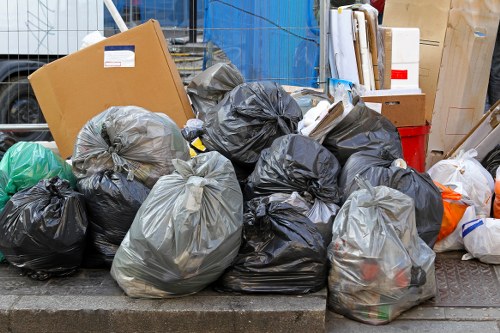 A collection of black and grey rubbish bags filled with waste materials, placed on a paved surface in an outdoor industrial or service area, with some bags tied at the top and others loosely gathered. The bags appear to contain general refuse, and they are positioned in front of a backdrop that includes a blue shipping container, wooden pallets, cardboard boxes, and other miscellaneous items. The scene suggests an active waste collection or clearance process, typical of professional rubbish removal services such as those offered by Office Clearance Brompton. The setting likely represents a temporary holding spot on a street or alley in Brompton, close to residential or commercial premises, awaiting removal or disposal, consistent with local rubbish collection practices in the area.