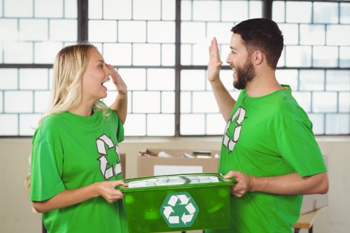 In a brightly lit indoor space, two individuals wearing green recycling t-shirts with white recycling symbols are engaging in a friendly gesture, possibly a high-five, while exchanging a green recycling bin that bears a white recycling icon on its side. The woman, with blonde hair, is on the left, and the man, with dark hair and a beard, is on the right. Behind them, large windows with metal grid frames allow natural light to fill the room. In the background, several cardboard boxes are visible, indicating an ongoing clearance or sorting activity, typical of waste management or rubbish collection operations in the Brompton area. The scene conveys a professional, environmentally conscious atmosphere aligned with rubbish removal services offered by Office Clearance Brompton, emphasizing recycling efforts and responsible waste handling. The textures of the recycling bin appear smooth and plastic-like, with the icon prominently displayed, while the clothing of both individuals looks breathable and suitable for manual work. The overall environment is clean, orderly, and focused on waste segregation and recycling in a commercial or warehouse setting.