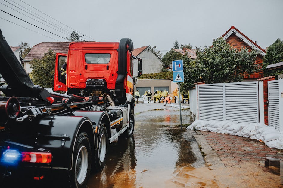 A group of workers dressed in high-visibility yellow and orange waterproof jackets and protective helmets are gathered on a residential street during daytime, actively engaged in the removal of flood-