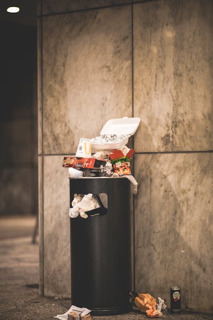 A middle-aged man with short dark hair, wearing a black T-shirt with white text and dark pants, is seen leaning over a cylindrical stainless steel rubbish bin on a paved outdoor walkway. His right han